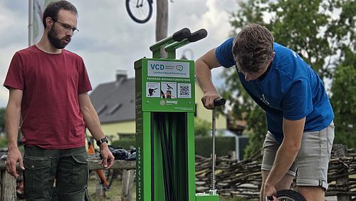 Zwei Personen stehen an einer grünen Fahrradreparatursäule; eine Person repariert ein Fahrrad, während die andere zusieht.
