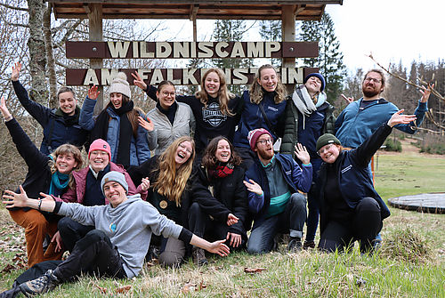 14 PraktikantInnen posieren freudig vor einem Holzschild mit der Aufschrift ,,Wildniscamp am Falkenstein" im Freien.
