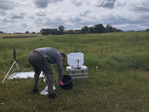 Eine Person mit abgewandtem, nach unten gerichtetem Gesicht beugt sich in einer Wiesenlandschaft über Kabel und einen schwarzen Behälter. Daneben stehen Teile der automatischen Messstation.