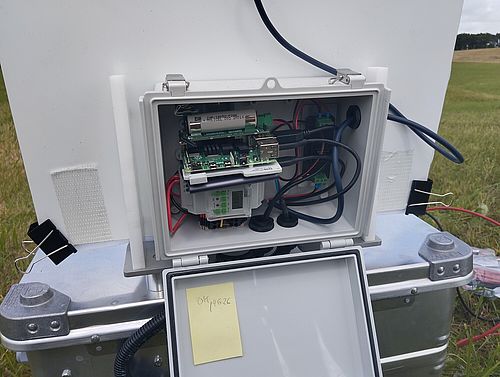 Nahaufnahme einer geöffneten weißen Steuerungsbox. Darin sind eine kleine Rechnerplatine, Kabel, Anschlüsse und weitere elektronische Bauteile zu sehen.