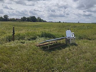 Weite Wiesenlandschaft unter wolkigem Himmel. Rechts im Vordergrund steht eine Messstation aus Metallkisten, einer weißen Insektenkamera und einem schräg montierten Solarmodul.