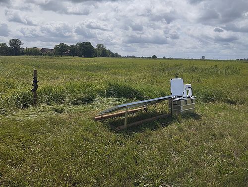 Weite Wiesenlandschaft unter wolkigem Himmel. Rechts im Vordergrund steht eine Messstation aus Metallkisten, einer weißen Insektenkamera und einem schräg montierten Solarmodul.
