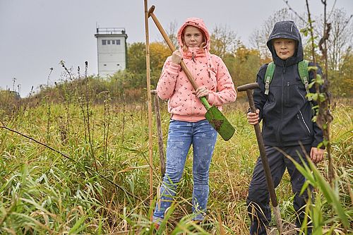 Zwei Schüler stehen, mit Spaten bewaffnet, vor einem alten Grenzturm im Feld.