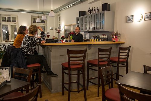 Zwei Frauen sitzen auf Hockern an der Bar, Barkeeper steht dahinter. 
