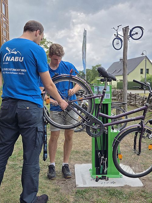Zwei Personen stehen an einer grünen Fahrradreparatursäule. Ein Fahrrad ist mit beiden Rädern nach oben an der Halterung befestigt.
